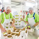 Drei Mitarbeitende in grünen T-Shirts und weißen Latzhosen stehen lachend in der Produktion und halten Lobetaler-Joghurt.
