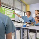 Vier Personen in OP-Kleidung stehen im Krankenhaus. Ein Mann zeigt mit ausgestrecktem Arm auf einen großen Bildschirm.