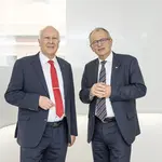 Herr Habenicht und Herr Lilie stehen gemeinsam in einem hellen, weißen Raum und tragen beide einen dunkelblauen Anzug.