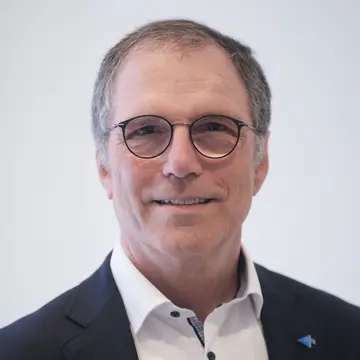 Herr Bernhard Schneider, lächelt. Er trägt eine Brille und ein weißes Hemd. 