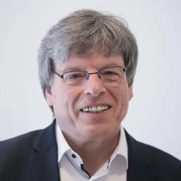 Herr Daub, lacht. Er trägt ein weißes Hemd und schwarzes Sakko. 