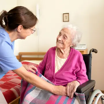 Das Bild zeigt eine Pflegerin (links), die sich einer Seniorin im Rollstuhl zuwendet und ihre Hände hält. Die ältere Frau lacht. 