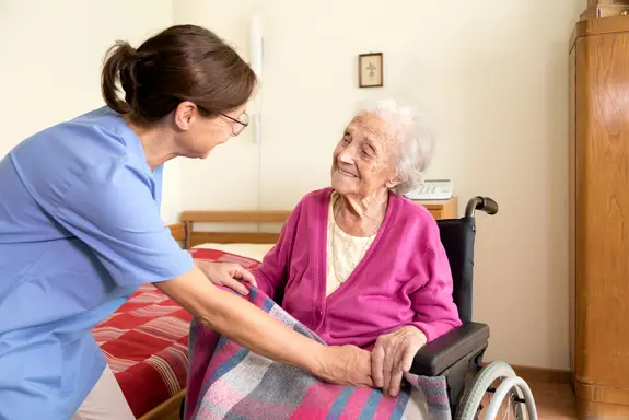 Das Bild zeigt eine Pflegerin (links), die sich einer Seniorin im Rollstuhl zuwendet und ihre Hände hält. Die ältere Frau lacht. 