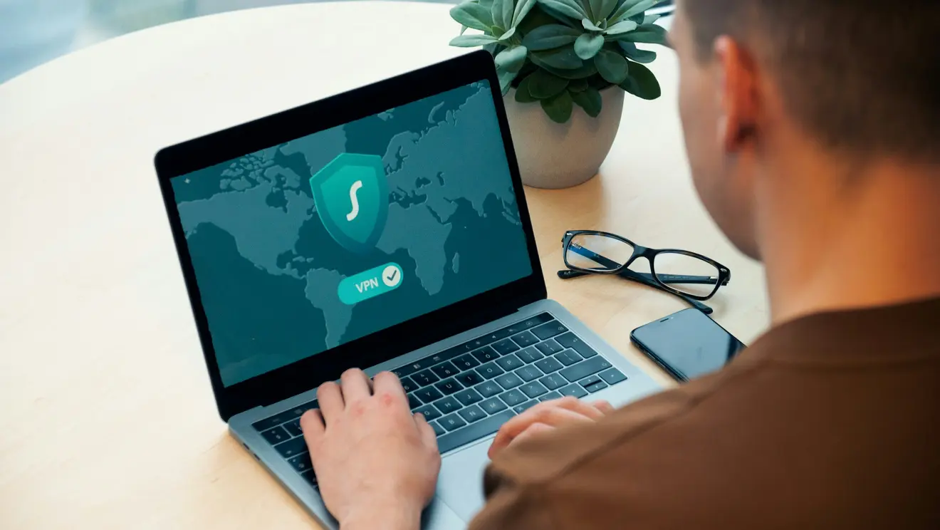 Ein Mann sitzt an einem Laptop. Ein Schutzsymbold auf dem Bildschirm steht für das Thema Cybersicherheit.