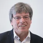 Herr Daub, lacht. Er trägt ein weißes Hemd und schwarzes Sakko. 