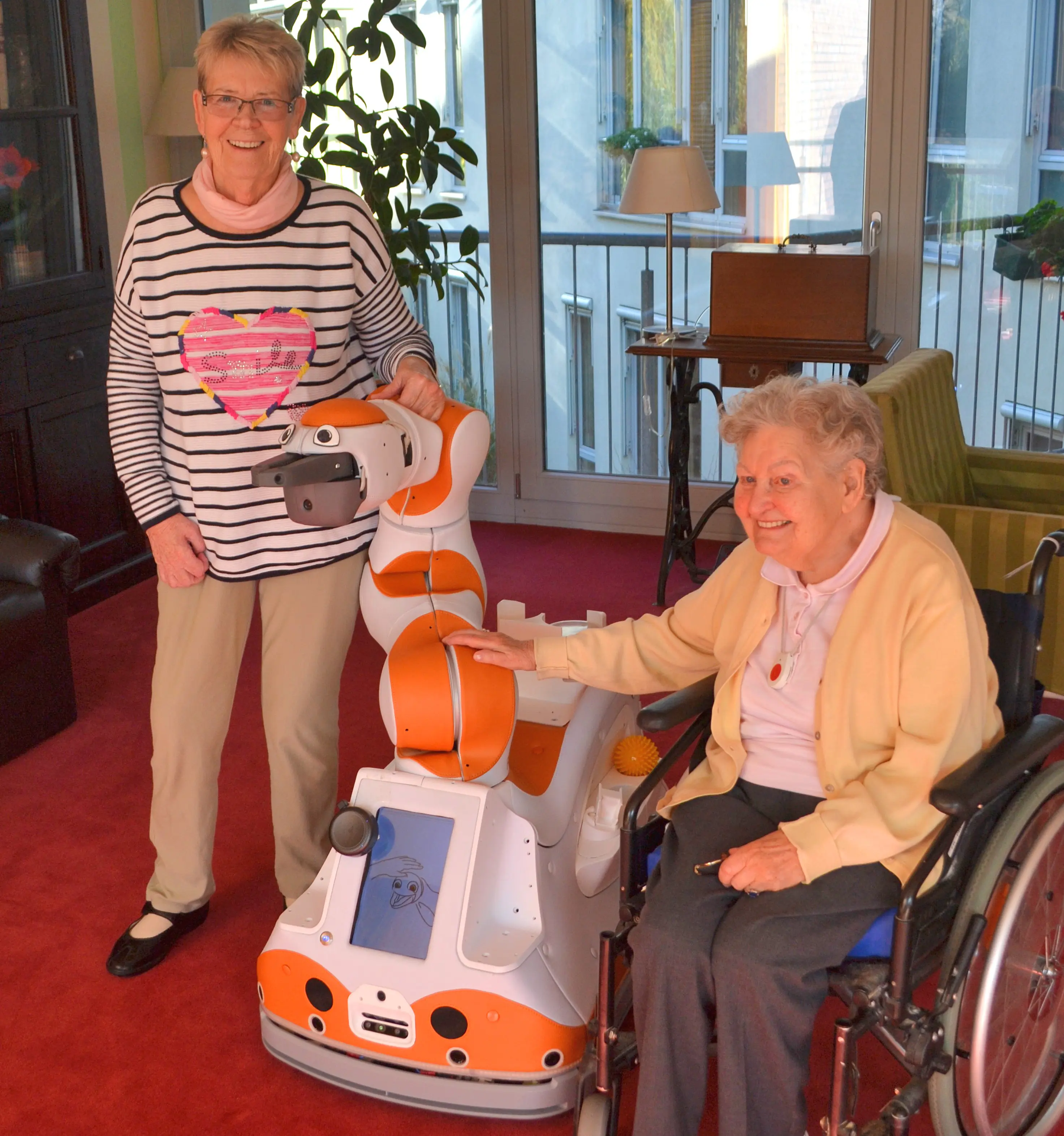 Zwei Seniorinnen stehen lachend neben Roboter LIO, der in der Mitte positioniert ist.