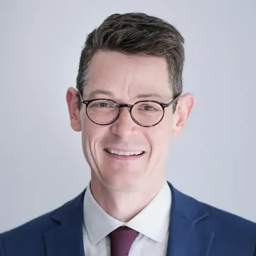 Dr. iur. Max Mälzer lacht. Er trägt Anzug, rote Krawatte und eine Brille. 