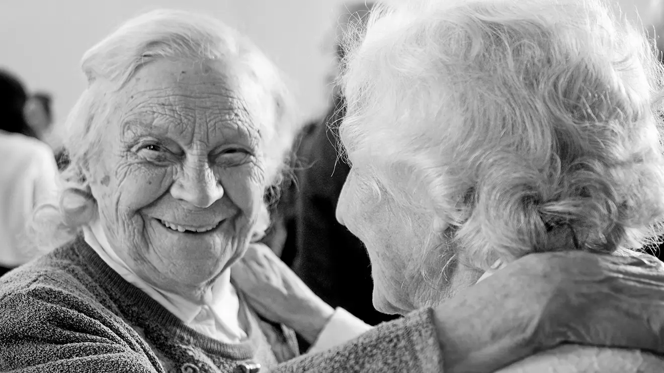 Das Bild in Graustufen zeigt zwei alte Frauen, die sich umarmen. Eine der Frauen blickt direkt in die Kamera und lacht.