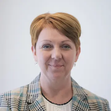 Frau Verena Hoelken trägt einen karierten Blazer, eine weiße Bluse und schlichte Ohrringe.