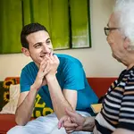 Ein junger Mann sitzt einer Seniorin gegenüber. Beide sind einander zugewandt und unterhalten sich, während der junge Mann lacht.