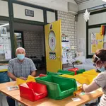 Das Bild zeigt zwei Personen, die in einer Werkstatt arbeiten. Sie sitzen an einem Tisch. Vor ihnen stehen verschiedene Arbeitsmaterialien und farbige Boxen.