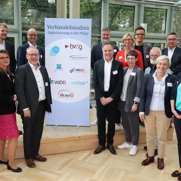 Das Bild zeigt 15 Personen. Sechs stehen links und 9 rechts. In der Mitte steht ein Banner mit der Aufschrift: Verbändebündnis Digitalisierung in der Pflege. Sie stehen in einem lichtdurchfluteten Raum. 