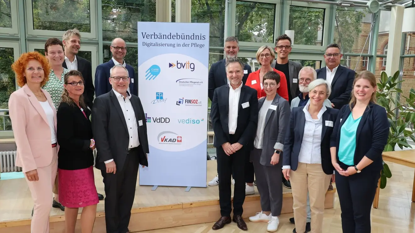 Das Bild zeigt 15 Personen. Sechs stehen links und 9 rechts. In der Mitte steht ein Banner mit der Aufschrift: Verbändebündnis Digitalisierung in der Pflege. Sie stehen in einem lichtdurchfluteten Raum.