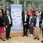 Das Bild zeigt 15 Personen. Sechs stehen links und 9 rechts. In der Mitte steht ein Banner mit der Aufschrift: Verbändebündnis Digitalisierung in der Pflege. Sie stehen in einem lichtdurchfluteten Raum.