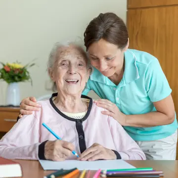 Eine Pflegerin beugt sich zu einer älteren Frau und berührt sie an den Schultern. Die ältere Frau lacht und hält einen Stift in der Hand. 