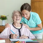 Eine Pflegerin beugt sich zu einer älteren Frau und berührt sie an den Schultern. Die ältere Frau lacht und hält einen Stift in der Hand. 