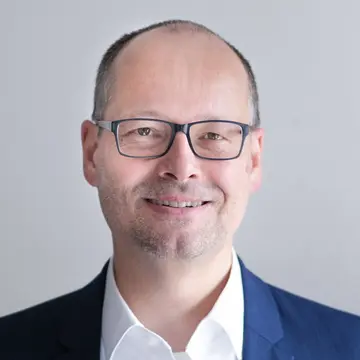 Herr Rolf Baumann lächelt, trägt eine eckige schwarze Brille, ein weißes Hemd und ein blaues Sakko.
