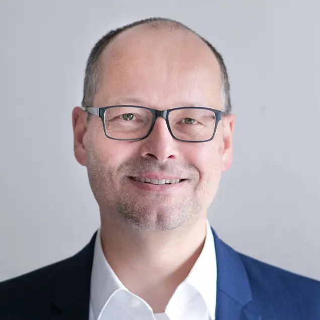 Herr Rolf Baumann lächelt, trägt eine eckige schwarze Brille, ein weißes Hemd und ein blaues Sakko.
