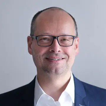 Herr Rolf Baumann lächelt, trägt eine eckige schwarze Brille, ein weißes Hemd und ein blaues Sakko.