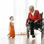 Ältere Frau im Rollstuhl beugt sich zu kleinem Roboter namens Navel, der links in orangefarbener Kleidung steht.