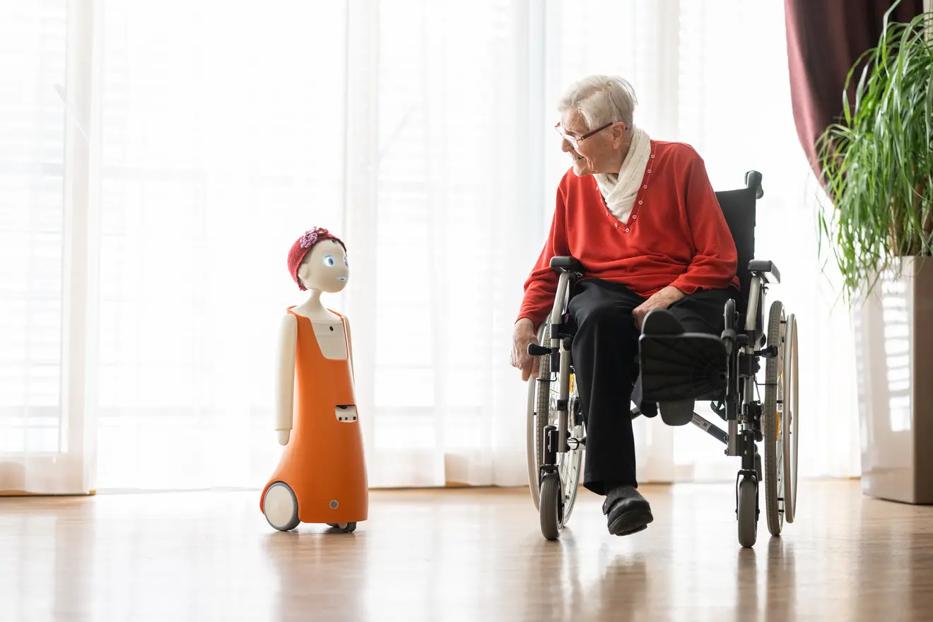 Ältere Frau im Rollstuhl beugt sich zu kleinem Roboter namens Navel, der links in orangefarbener Kleidung steht.