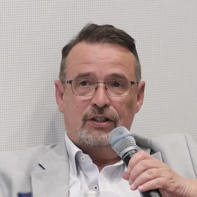 Herr Dopheide, hält ein Mikrofon in der Hand. Er trägt helle, professionelle Berufskleidung, eine Brille und Bart. 