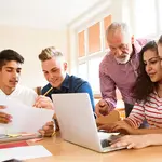 Vier junge Menschen sitzen am Tisch mit Laptops und Schreibunterlagen. Der Lehrer beugt sich vor und zeigt etwas am Laptop.