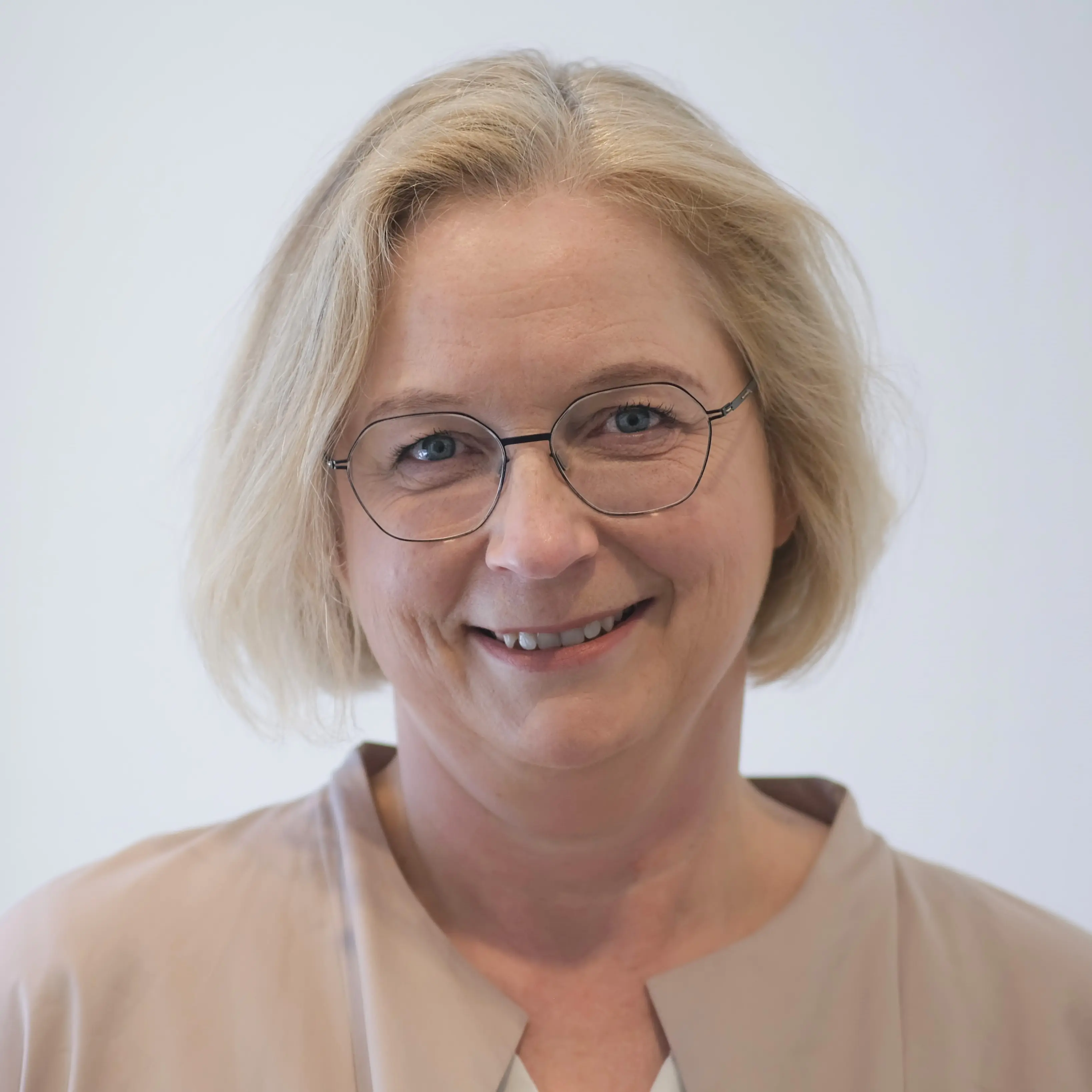 Frau Hannemann ist beigefarben gekleidet und trägt eine runde Brille. Ihre Haare sind blond und kinnlang.  