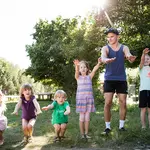 Fünf Kinder hüpfen fröhlich lachend mit einem FSJler gemeinsam im Garten. Die Sonne scheint. 