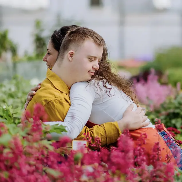 Eine junge Frau umarmt einen Jungen mit Trisomie 21 in einem Blumenfeld.