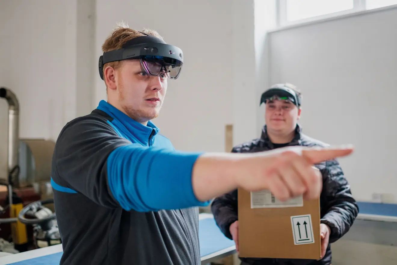 Zwei Männer tragen Augmented-Reality-Brillen. Der vordere Mann zeigt mit dem Finger in eine Richtung, während der hintere Mann einen Karton hält.