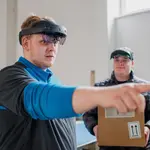 Zwei Männer tragen Augmented-Reality-Brillen. Der vordere Mann zeigt mit dem Finger in eine Richtung, während der hintere Mann einen Karton hält.