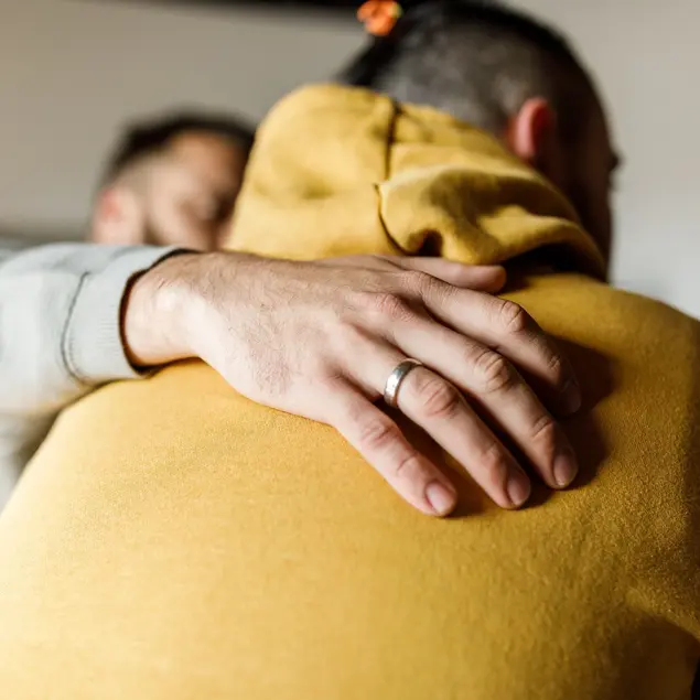 Eine Person legt beruhigend die Hand auf den Rücken einer anderen Person in einem gelben Pullover.