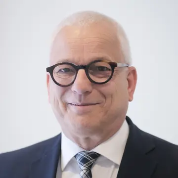 Herr Hubertus Jäger trägt eine schwarze, markante und runde Brille. Er lächelt und trägt einen Anzug. 